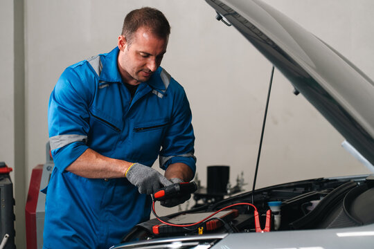 And keywordsMechanic man blue coverall car engine battery test electrical repair garage focused expression while inspecting vehicle battery and wiring during routine automotive maintenance