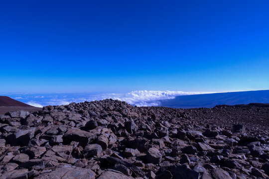 Field of broken lava rock on the summit plateau of Mauna Kea above a closed sea of clouds with the shield of Mauna Loa on the horizon