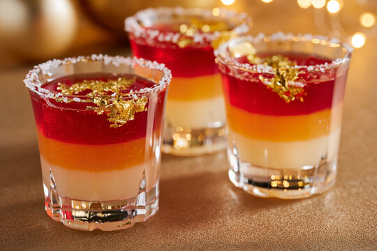 Colorful layered jelly shots with red, orange, and yellow layers topped with gold flakes in clear glasses on a festive table setting