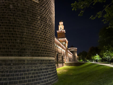 Sforza Castle glows against the night sky in Milan, its brick towers illuminated as shadowy walls frame a peaceful garden.