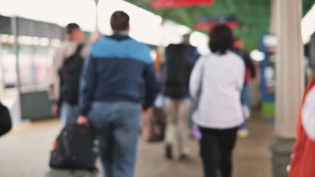 Individuals walk through a busy train station, carrying luggage and backpacks. The scene shows people in various outfits moving along the platform, defocus
