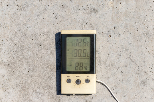 Digital thermometer and hygrometer with a sensor wire resting against a rough concrete wall.