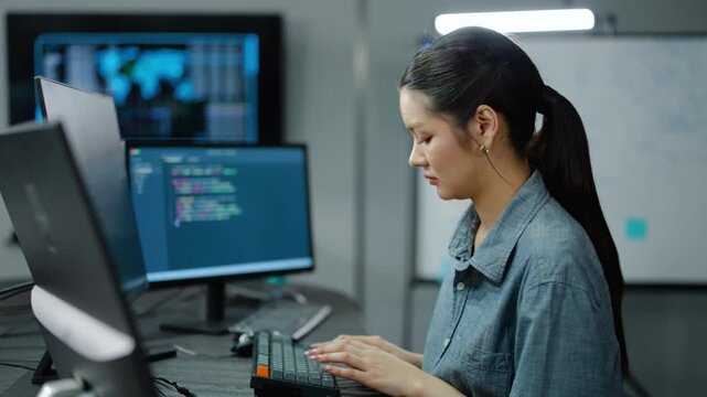 Exhausted Asian female programmer coding late at night in a dark office with multiple monitors. Tired IT woman working on cybersecurity systems. Overworked tech expert typing code at her desk.