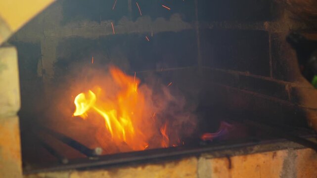 Person dangerously pouring flammable lighter fluid onto an open fire, causing a large flare-up and smoke - Hand squeezing liquid accelerant bottle to ignite a hot fire in a brick barbecue pit