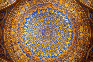 Symmetrical view of an ornate Islamic dome ceiling in Samarkand, Uzbekistan, featuring a vivid blue and gold floral medallion surrounded by intricate muqarnas, geometric star motifs © pploylp