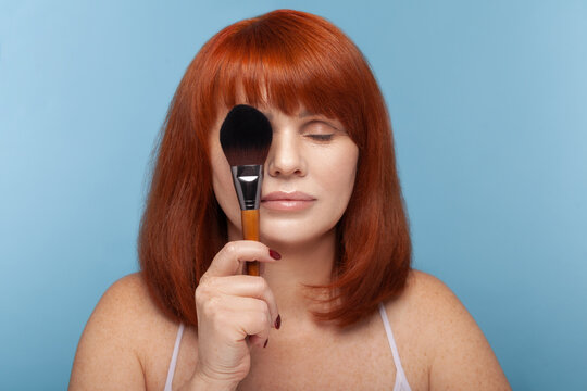 Calm mature redhead woman holding large black makeup brush in front of her face with closed eyes during beauty visage process isolated on blue background