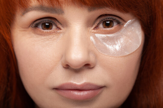 Close up portrait of middle aged redhead woman looking directly at camera while wearing an anti-wrinkle hydrogel eye patch isolated on blue background