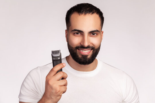 Smiling man holding electric shaver for grooming posing with professional cordless beard trimmer wearing white t-shirt isolated over gray background