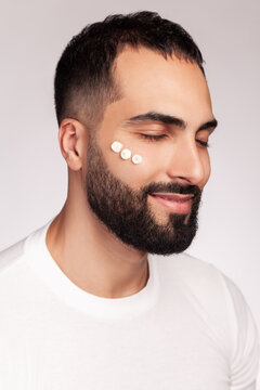 Bearded man applying three white cream drops on cheek skin for daily beauty routine grooming procedure holding eyes closed isolated over gray background