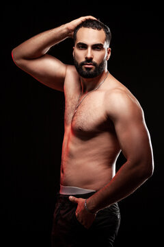 Athletic man holding hand on head showing powerful triceps and well defined pectoral muscles wearing dark pants and silver jewelry isolated over black background