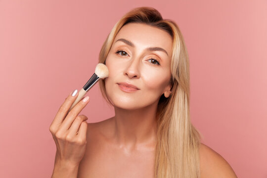 Beautiful lady applying makeup with professional brush enhancing cheekbones and facial features daily cosmetic routine isolated on pink background