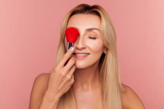 Playful woman covering eye with large red makeup brush smiling and enjoying daily cosmetic procedure at home isolated on pink background