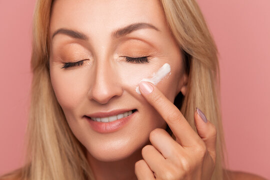 Mature blonde woman smiling gently with closed eyes while applying anti-aging moisturizer with her finger on cheek isolated on pink background