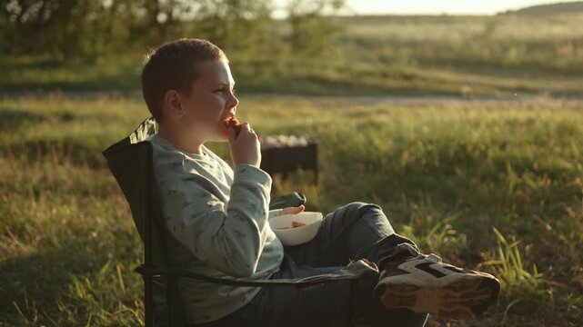 Caucasian child sits in folding chair at picnic on edge of forest, nibbling on food at sunset. Portrait of relaxed boy enjoying rest and view during camping trip.