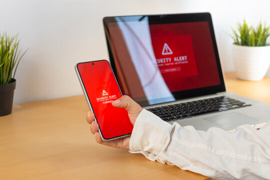 Man holding smartphone with security alert and laptop in background