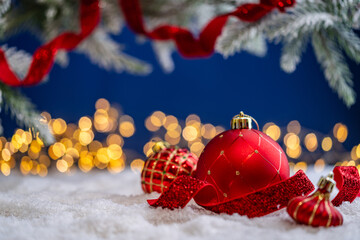 Naklejka premium A Christmas background featuring red baubles with gold quilted patterns and glitter ribbon on snow, framed by frosted pine, set against a deep blue backdrop with twinkling lights and copy space.