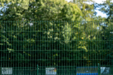 Zielone metalowe ogrodzenie panelowe z siatki na tle gęstych rozmytych drzew w słoneczny dzień © mbugajsky