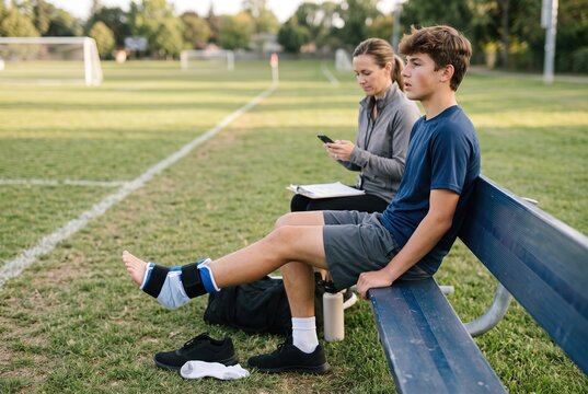 A young boy rests on a bench with an injured ankle wrapped in a bandage during soccer practice. His parent looks at their phone while watching him. The setting is a soccer field during the afternoon