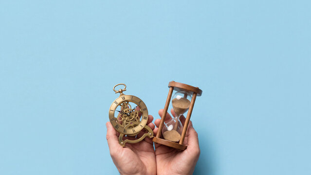 Hourglass and vintage clock held in hands represent time management concepts on a solid blue background.