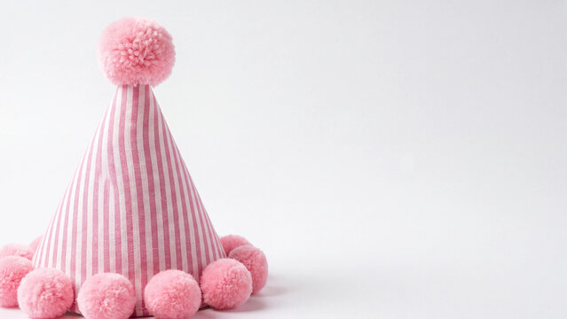 Party hat with pink stripes and pom poms standing upright on a simple white studio surface.