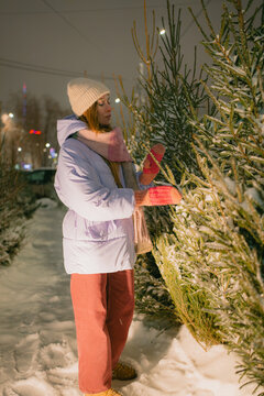Woman in warm beanie, scarf and mittens reaches to touch a snow-dusted christmas tree at a nighttime outdoor market, enjoying the festive, wintry holiday shopping atmosphere