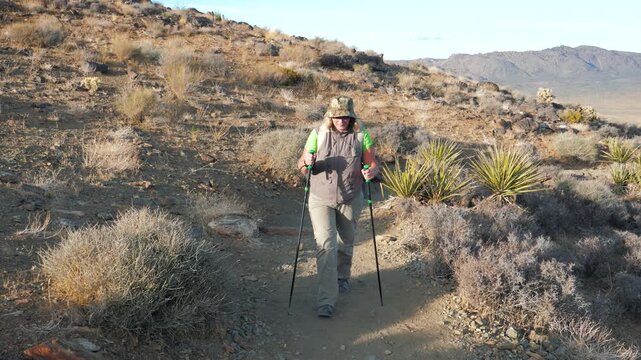 Mature woman with backpack and trekking poles actively hiking along narrow dirt path in rugged, arid hills landscape enjoying an adventurous journey through dry sunny wilderness. Hiking activities