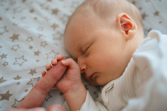 Newborn baby sleeping soundly in a bed with star patterns, gently holding an adult's finger, illustrating the close bond, innocence, and care within a family, marking the beginning of life