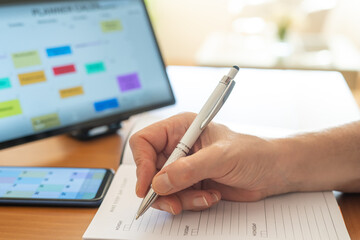 Person's hand writing daily tasks and appointments in a paper planner, cross-referencing with a...