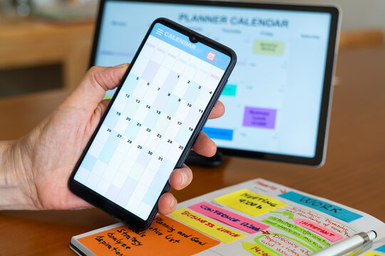 Hand holding a smartphone displaying a calendar app, coordinating tasks and appointments with a planner calendar on a tablet and a handwritten agenda for efficient time management