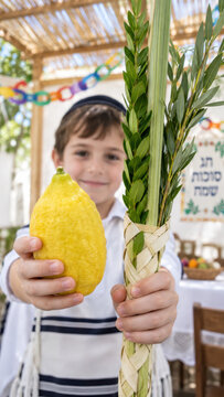 Boy in tallit presenting etrog plus lulav, jewish child standing inside decorated sukkah for holiday observance, family celebration article for seasonal faith gathering
