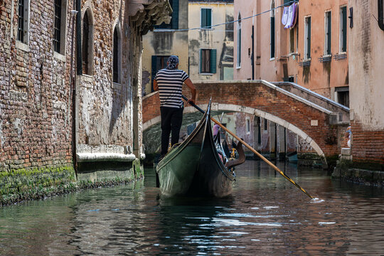 A gondolier navigating a narrow canal in Venice, Italy, passing under a small brick arch bridge