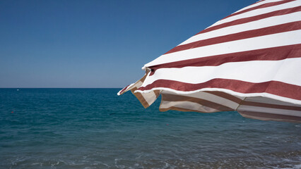 A striking view of a striped beach umbrella overlooking the tranquil blue sea under a clear sky. © Azur