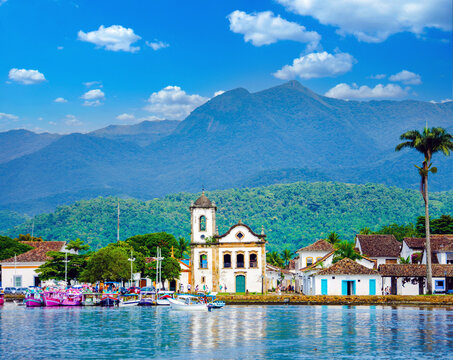 Scenic coastal view of historic Paraty, Brazil, with a boat, Santa Rita church, and lush mountains under a bright blue sky.