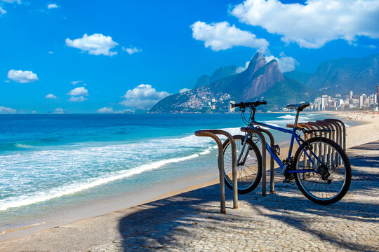 Bicycle parked on the iconic mosaic promenade of Ipanema beach in Rio de Janeiro, with the Dois Irmaos mountains and blue ocean in the background on a sunny day.