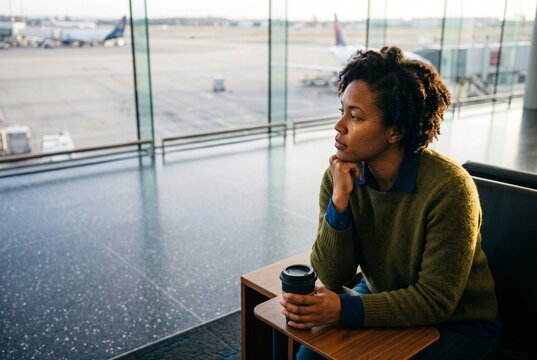 Thoughtful Black woman sitting in an airport terminal lounge holding a coffee cup while looking out the window at airplanes
