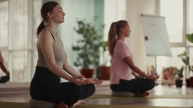 Women meditating in lotus pose at a yoga class in a bright room.