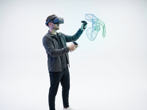 Young man using VR headset and controllers interacting with 3D wireframe hologram in white space