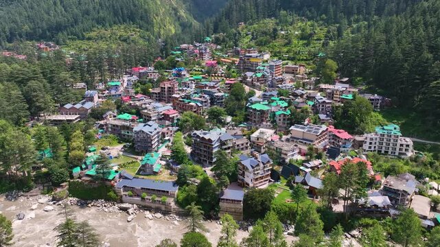 High angle drone reveal of the sprawling alpine town of Kasol nestled deeply within lush green Deodar forests, a premier destination for Kheerganga trekkers.