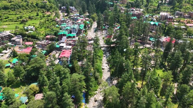 High angle drone tracking shot looking directly down at the colorful tin roofs and winding roads of Kasol town beside the fast-flowing Parvati River.