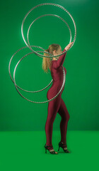 Side view full shot of a young circus gymnast in a red leotard standing on a green background, spinning metal hoops around her arms . © kinomaster