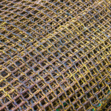 A detailed close-up of piled, textured yellowish-brown commercial fishing nets on a deck, featuring complex mesh patterns, knots, and a visible yellow hardware ring.