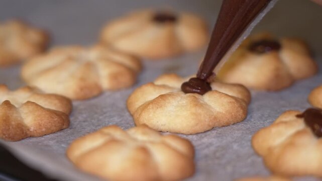A baker uses a piping bag to add chocolate icing to freshly baked cookies arranged on parchment paper, showcasing the intricate decoration process in a cozy kitchen setting