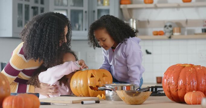 African American mom with girls steadying child carving pumpkin on island, scooping seeds for fall