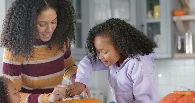 African American mom guiding older daughter carving pumpkin at counter, scooping into metal bowl