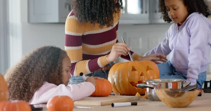 After marking pumpkin, African American mom guiding girls scooping pulp at kitchen island teaching
