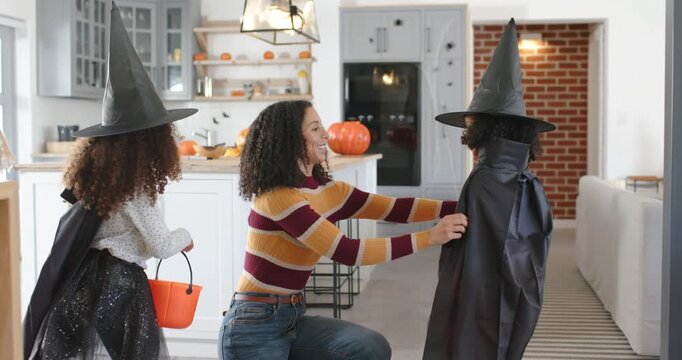African American mom in striped top, jeans fixing costumes, giving candy from pumpkin pail at home