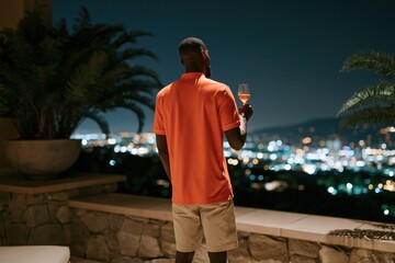 Obraz premium Man in orange polo holding a wine glass on a terrace overlooking a lit city skyline at night for luxury lifestyle wallpaper and rooftop background design