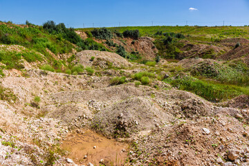Obraz premium Open limestone quarry with heaps of crushed stone, barren textured terrain, and overgrown greenery under a clear blue sky, showing industrial mining remnants in a natural setting