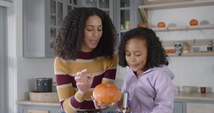 African American mom and daughter holding pumpkin as child reaching, drawing with marker in kitchen