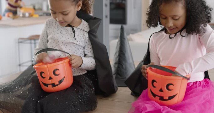 African American girls in capes sitting at kitchen island opening pumpkin pails to inspect candy